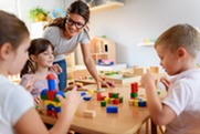 Woman at classroom desk with children playing and learning.