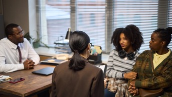 Middle aged Black man sitting at desk talking with middle aged Black woman, teenage girl and young adult woman in office