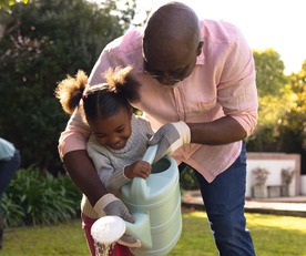 Happy senior african american man and his granddaughter watering plants in sunny garden