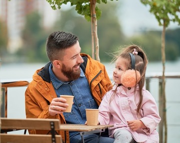 Man holding cup of coffee and smiling while spending time with child in outdoor cafe.