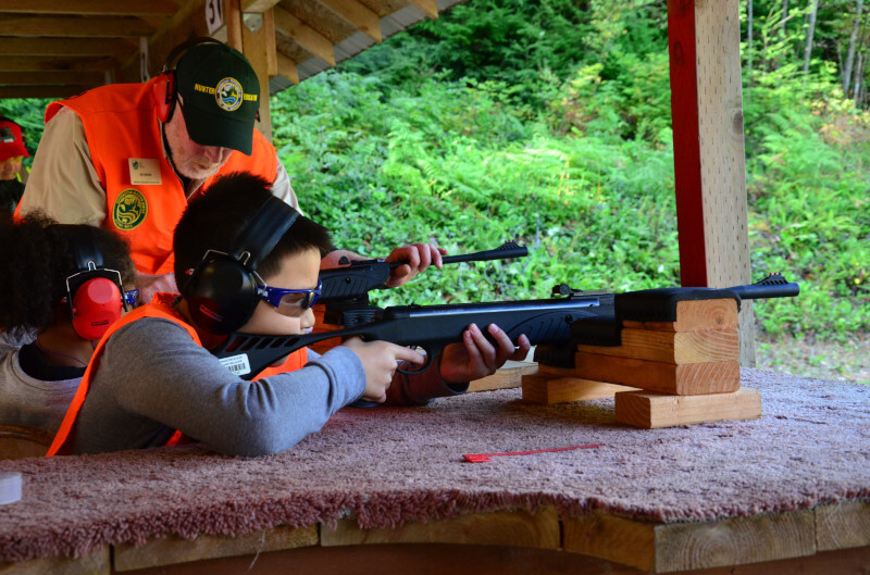 Two young students practicing at a shooting range, with a Hunter Education instructor nearby giving direction