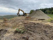 An excavator works near a shoreline alongside a pile of gravel