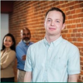 Teen worker stands in foreground looking forward at the camera as two adults in the background look at the teen 