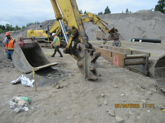 A bucket from an excavator lays next to an excavator arm. A trench with a trench box is nearby and another excavator is in the background.