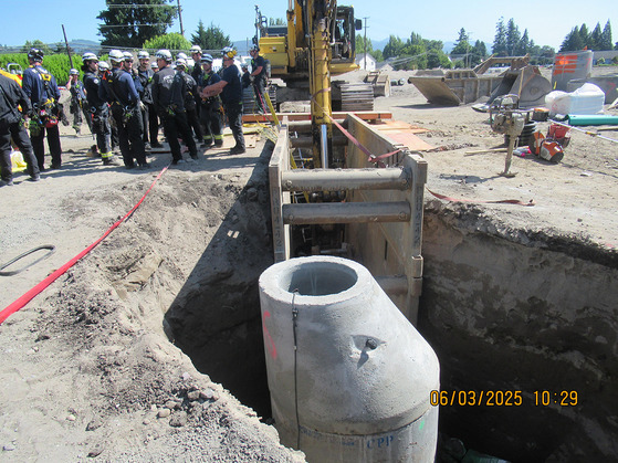 An upright cement sewer manhole is inside a deep trench. A trench box is next to it with an excavator arm inside. 