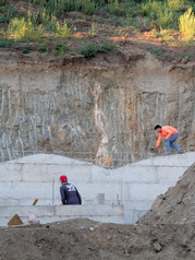 A worker is between concrete foundation blocks and next to wall of dirt. Another worker is on top of the concrete foundation blocks.