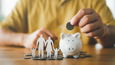 A person putting a coin into a piggy bank beside a paper family cutout.