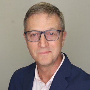 Headshot of Mark Harniss wearing glasses and a navy blazer against a light gray background.