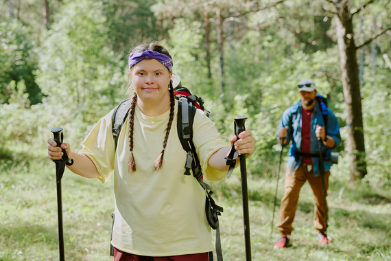 A young woman hikes on a wooded trail using trekking poles, with another hiker behind her.