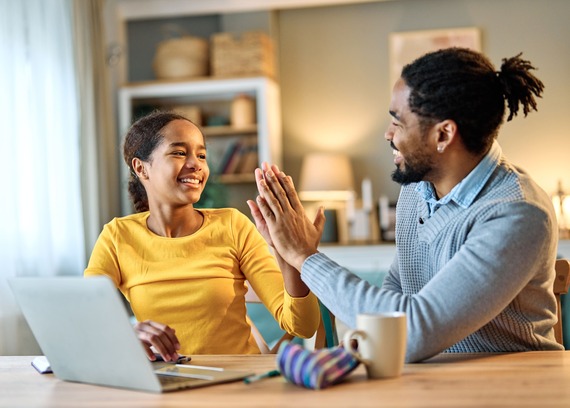 Father and daughter high five while sitting in front of a laptop computer against a blurred background.