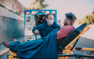 Male paramedic wearing mask talking to male patient on stretcher in front of ambulance 