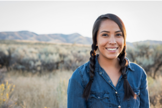 Young Native American woman standing outside with mountains behind her.