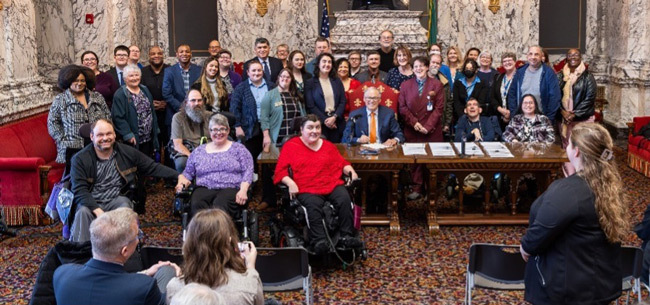 A large group of people stand and sit in wheelchairs gathered around Governor Inslee signing a piece of paper in front of a white marble walled room