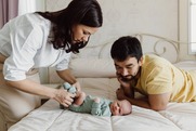 A mother puts stands in front of her newborn baby and puts socks on their feet while the new father watches and smiles down at the baby.