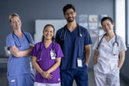 Four smiling nurses of different genders and ethnicities standing together in their uniforms of various colors. 