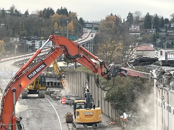 A large excavator arm removes a section of the old Montlake Boulevard overpass, looking west toward I-5.