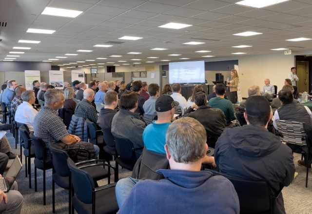 Photo shows a large crowd of seated people sitting before Program staff giving a presentation with a PowerPoint screen