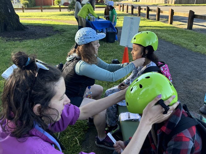 two adults help two students get fitted for bike helmets in a park 