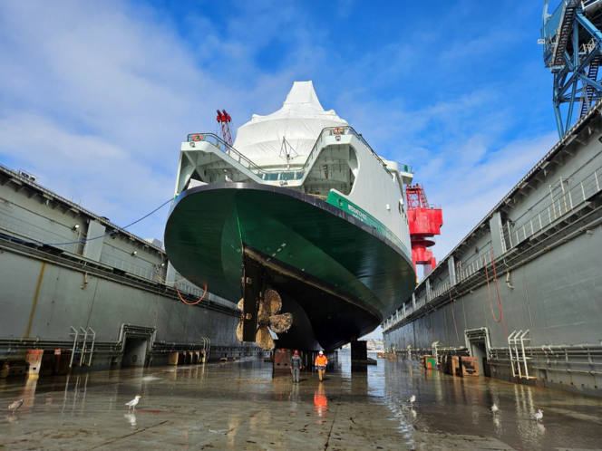 Large ship in dry dock with green hull and white covering, viewed from the stern