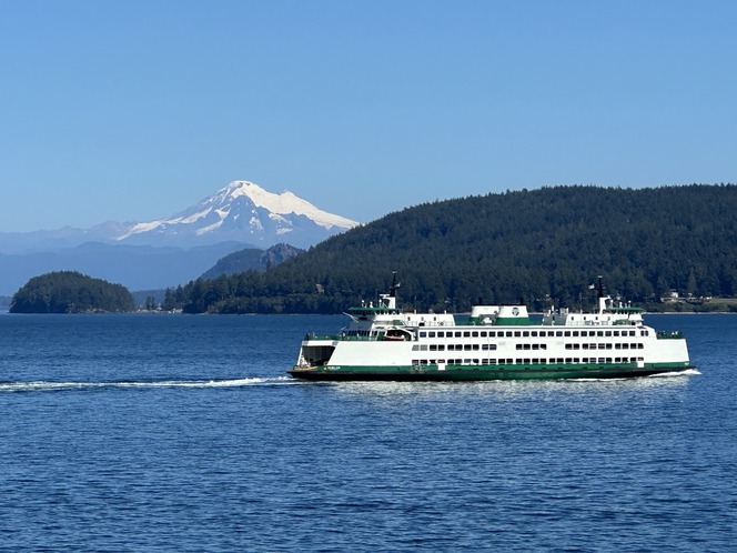 A ferry on blue water with a snow-capped mountain and forested hills in the background