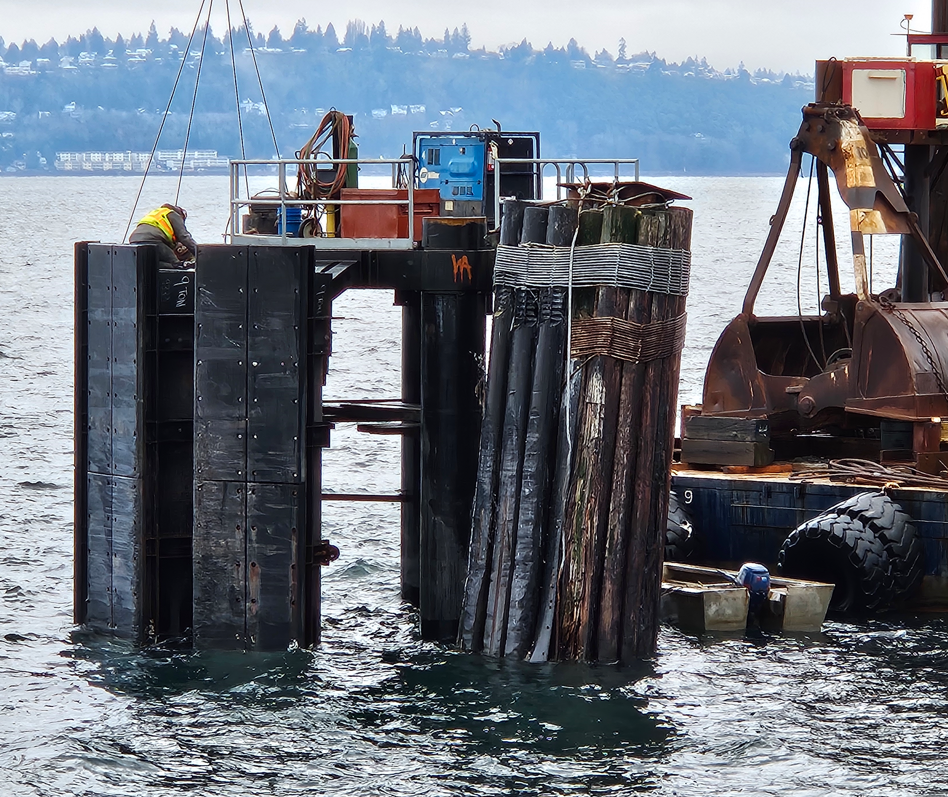 A sturdy metal structure partially submerged in water next to a bundle of logs and a barge with a crane