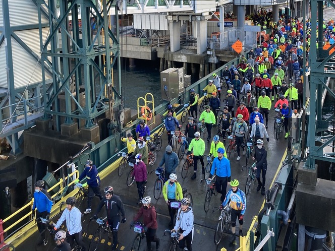 A large group of cyclists wearing colorful jackets walking with their bikes on a dock onto a ferry