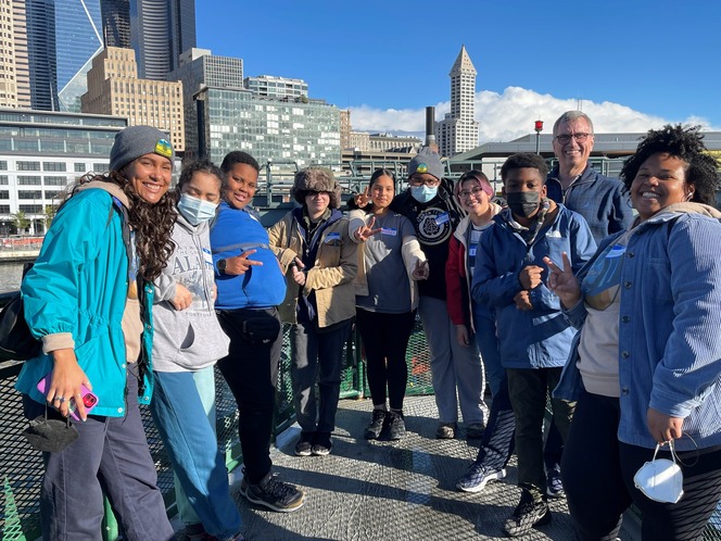 A diverse group of young adults posing together outdoors on a ferry with a city skyline in the background