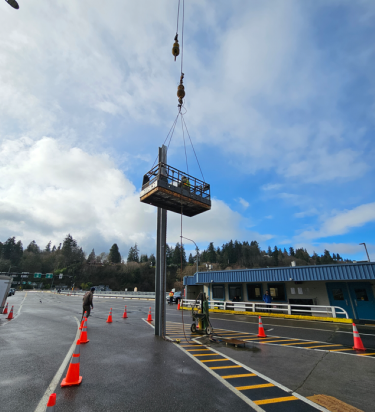 A construction platform suspended by a crane at Vashon terminal, with workers on the platform and traffic cones on the ground