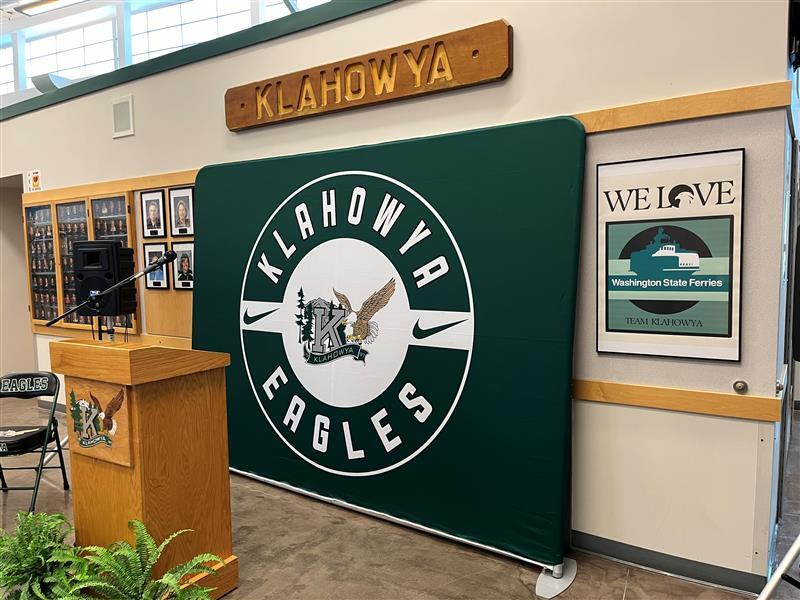 Indoor setting with a green backdrop featuring "KLAHOWYA EAGLES" emblem, a boat's nameplate "KLAHOWYA" and a wooden podium
