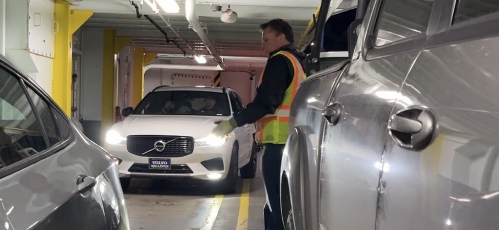 A person in a high-visibility vest directing a white Volvo with headlights on onto the car deck of a ferry