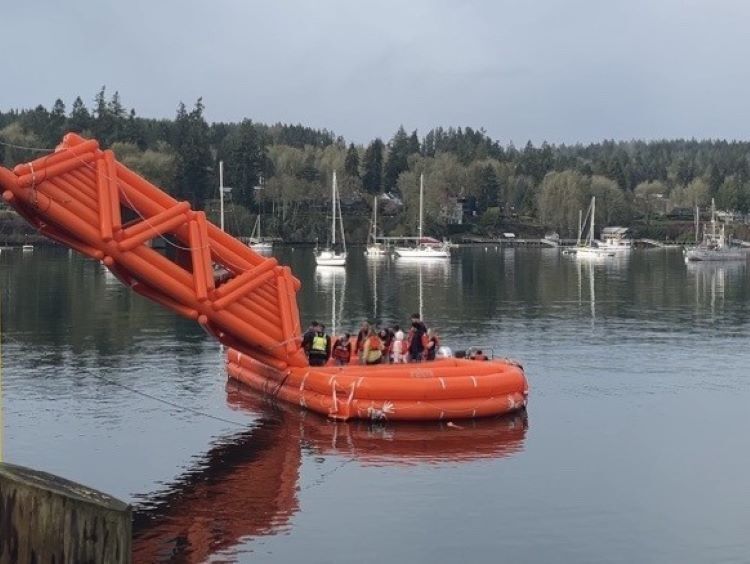 A group of people on an orange inflatable structure on water with sailboats and a tree-lined shore in the background