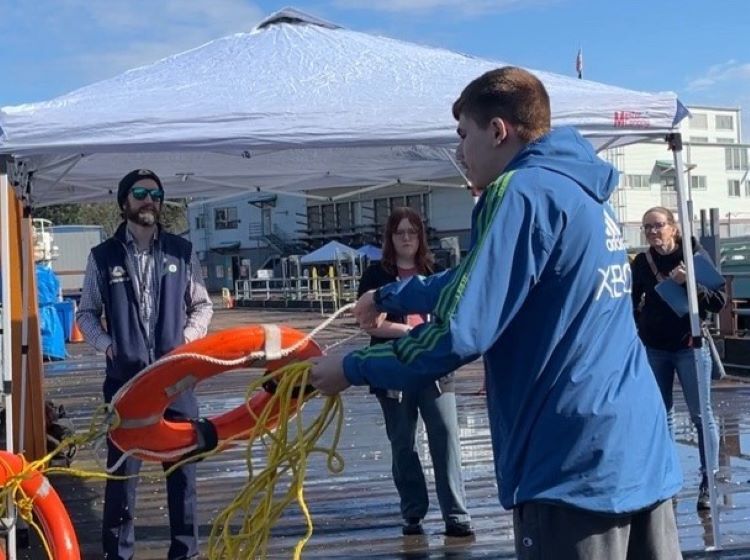 A person in a blue jacket throws a life ring with yellow ropes on a dock, under a white canopy tent