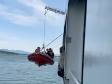 An inflatable rescue boat is suspended by a crane from the side of a larger vessel on a body of water