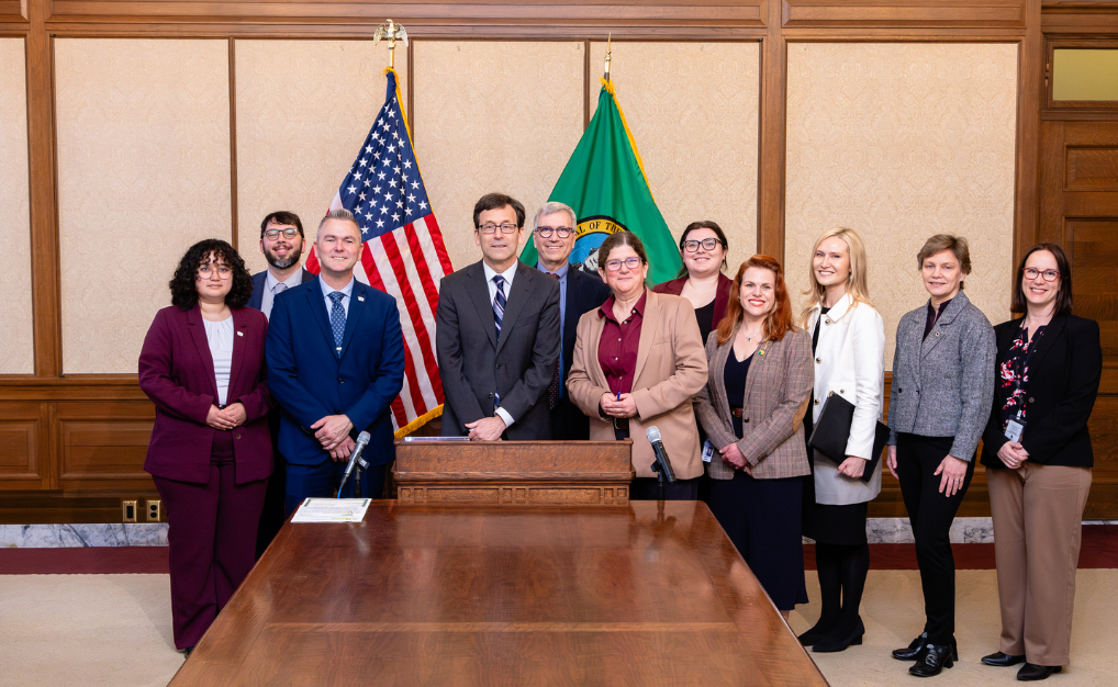 Ten people in business attire standing behind a table, with U.S. and state flags in the background