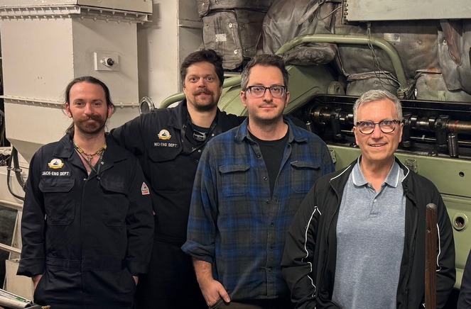 Four people in an engine room with large machinery in the background