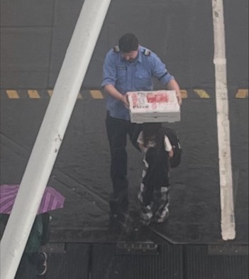 A person in a blue uniform uses pizza boxes to a protect a child from rain while they walk onto a ferry
