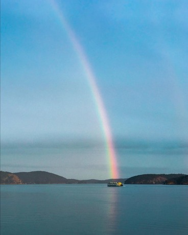 A ferry on a large body of water under a rainbow and blue sky with hills in the background