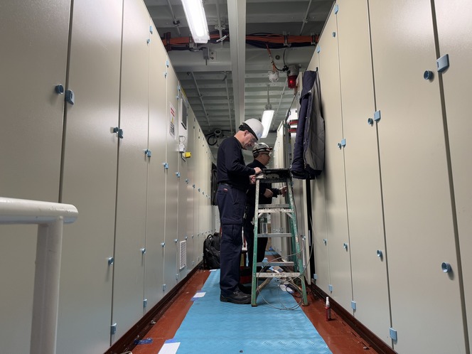 Two people in work attire and helmets in a corridor with walls of batteries on each side