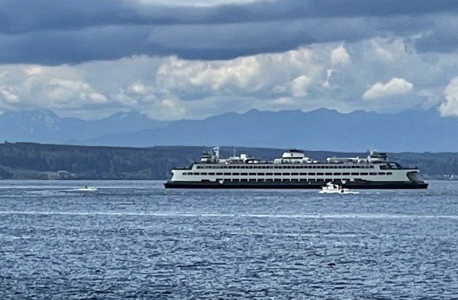 A ferry on a large body of water with other small boats nearby and mountains in the background under a cloudy sky