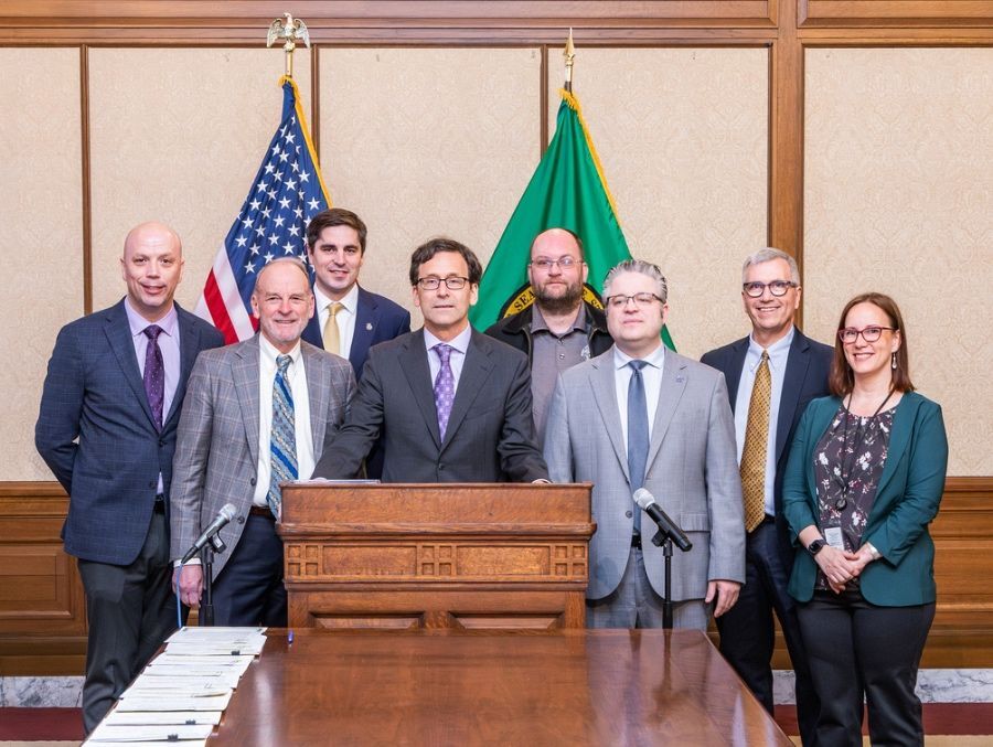 A group of eight people in business attire stand behind a wooden podium with microphones in a room featuring U.S. and Washington state flags.