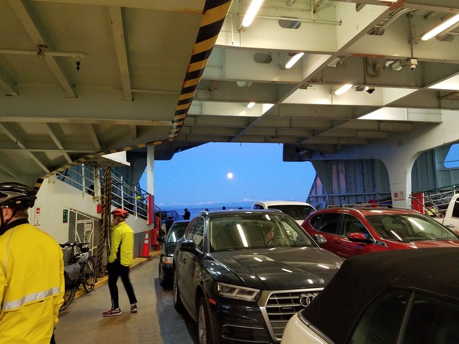 Inside a ferry with parked cars and bicycles, two people in yellow jackets walking, and a full moon visible through an opening in the background.