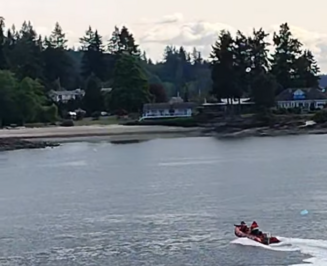 A rescue boat with two people on a calm body of water with an overturned blue kayak nearby with a tree-lined shoreline and houses in the background.