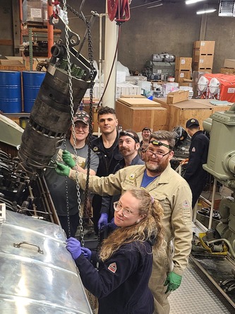 A group of seven people in a workshop, gathered around a suspended mechanical component.