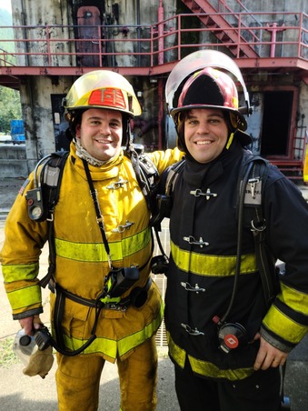 Two people in firefighting gear smiling in front of a concrete structure.