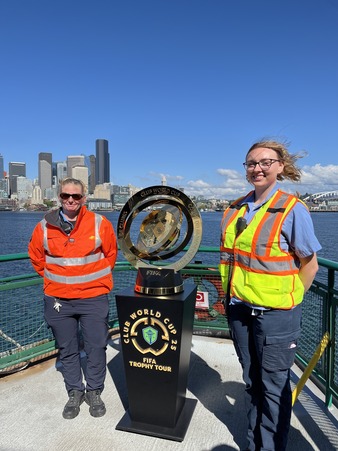 Two people in high-visibility clothing stand beside a gold and silver trophy on a ferry deck with a city skyline in the background.