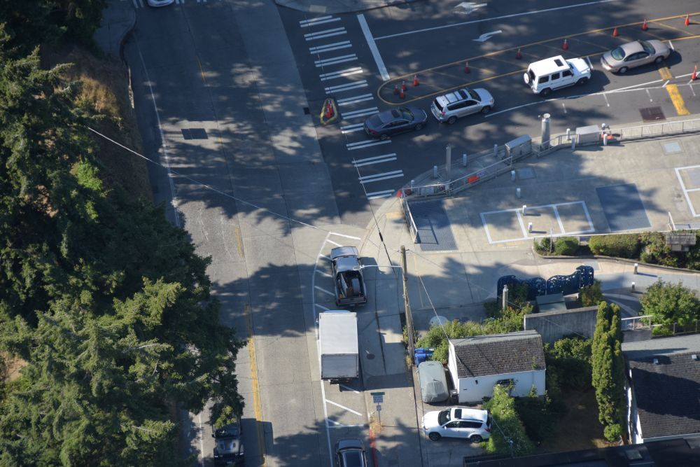 Aerial view of a road intersection with cars, orange cones, large tree, and nearby buildings.