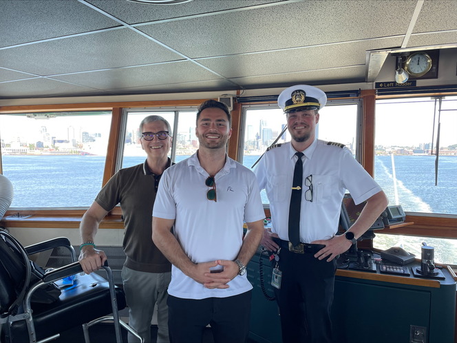 Three people standing in a ship's bridge with a harbor and city skyline visible through large windows.