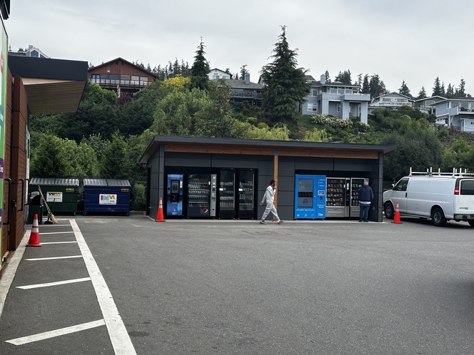 Outdoor scene with vending machines, dumpsters, and hillside houses in the background.