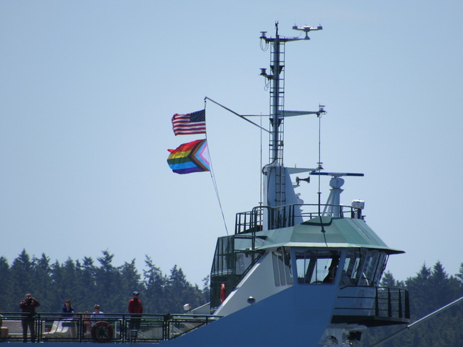 Ferry bridge with American and  Pride flags, radar tower, people on deck and trees in the background.