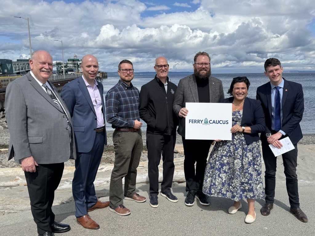 Group of seven people standing together, one holding a sign reading "Ferry Caucus" near a ferry dock.